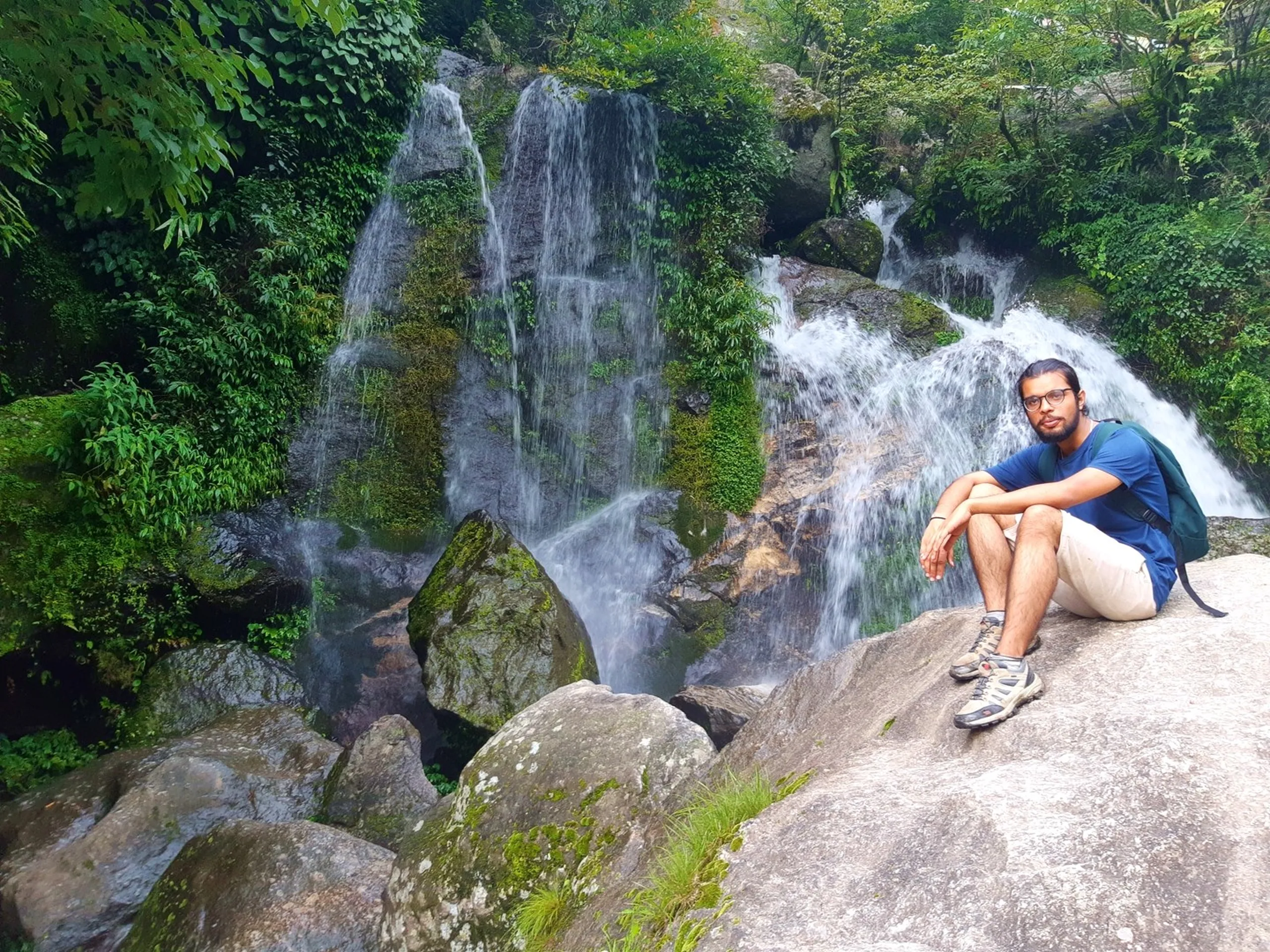Young male hiker wearing glasses and blue t-shirt with green backpack sitting relaxed on a large granite boulder directly beside a stunning multi-stream waterfall cascading down moss-covered green rocks surrounded by lush jungle - Sundarijal waterfall hiker Nepal day trek Kathmandu waterfall photography
