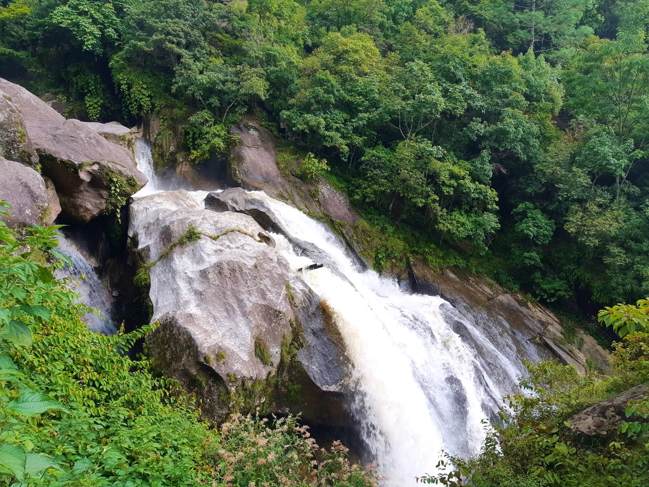 Powerful wide waterfall gushing forcefully over a massive smooth granite rock face at high speed, white foamy water rushing diagonally down the steep slope into a pool below, framed entirely by dense dark green monsoon forest - Sundarijal main waterfall Nepal monsoon heavy flow best waterfalls near Kathmandu