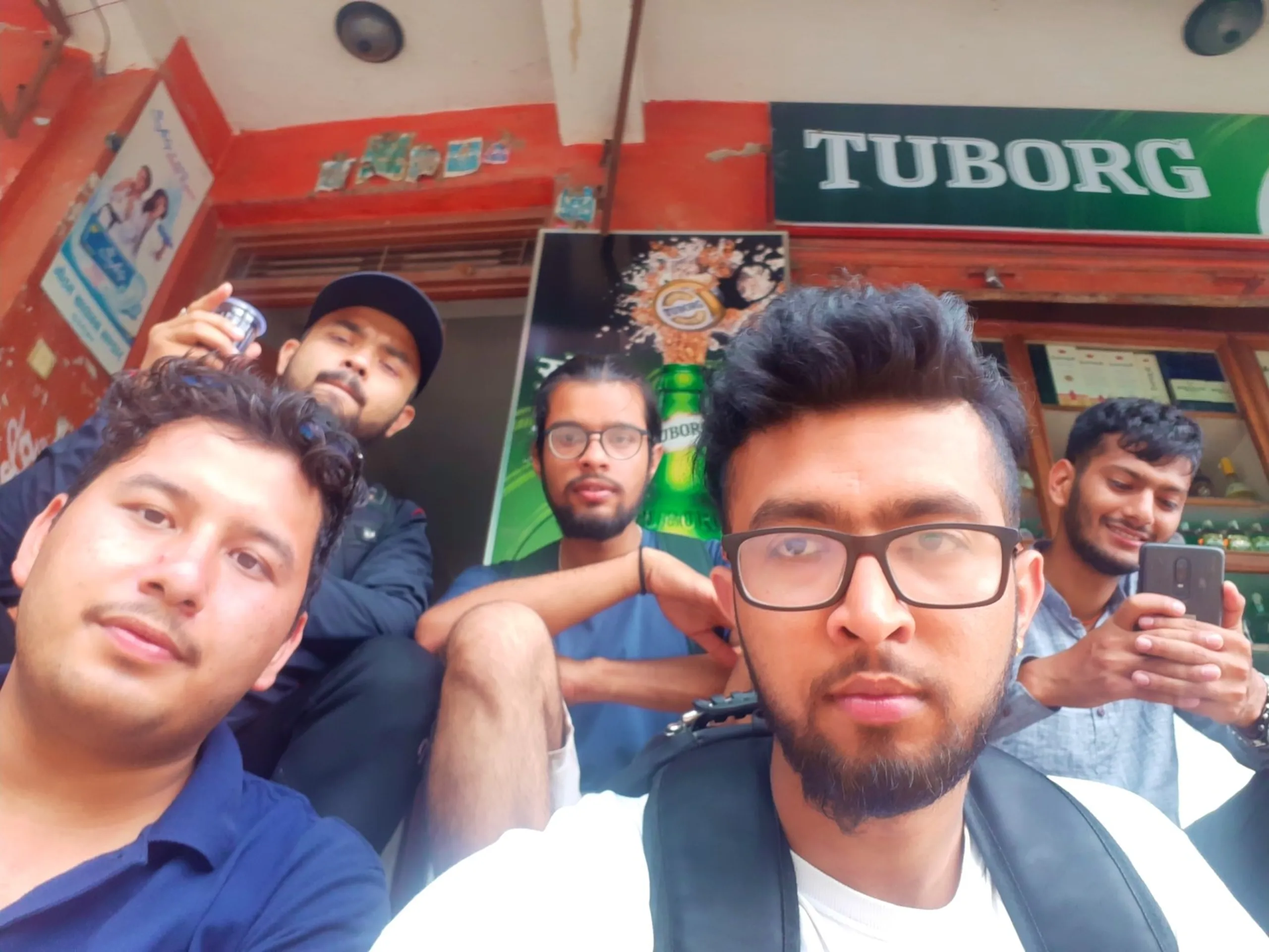 Fun wide-angle close-up selfie of five young Nepali male friends posing casually in front of a small local shop with a prominent green and white Tuborg beer signboard, one friend holding a drink, another on his phone, all with relaxed confident expressions - Nepal friends travel group Kathmandu local shop post-trek hangout
