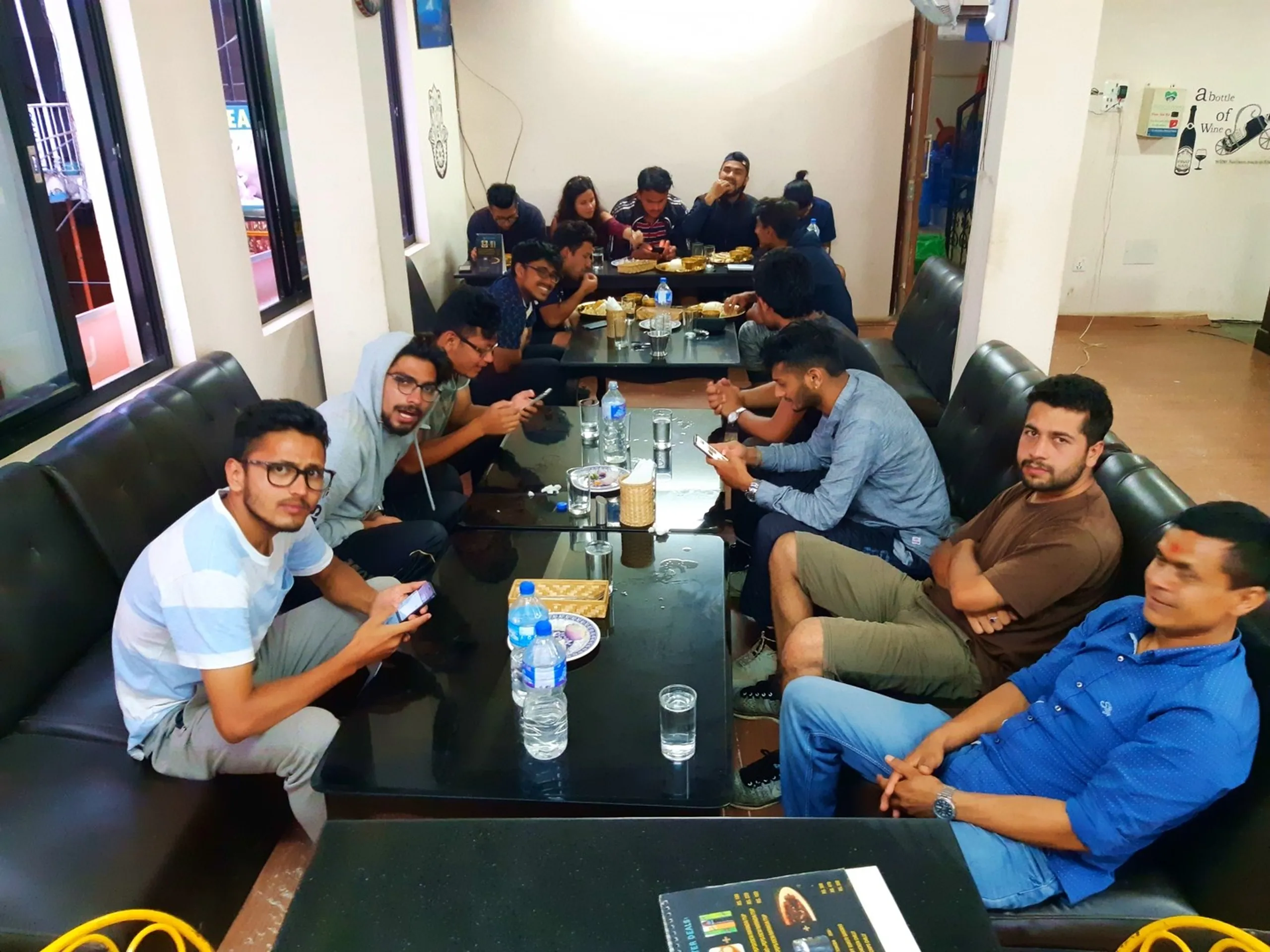 Large group of around fifteen young Nepali hikers and friends relaxing together inside a casual restaurant after a trek, seated on black leather sofas around dark glass tables with water bottles, food plates, and phones scattered across, warm indoor lighting, lively post-hike atmosphere - Nepal trekking group friends dinner Kathmandu restaurant post-hike meal hangout