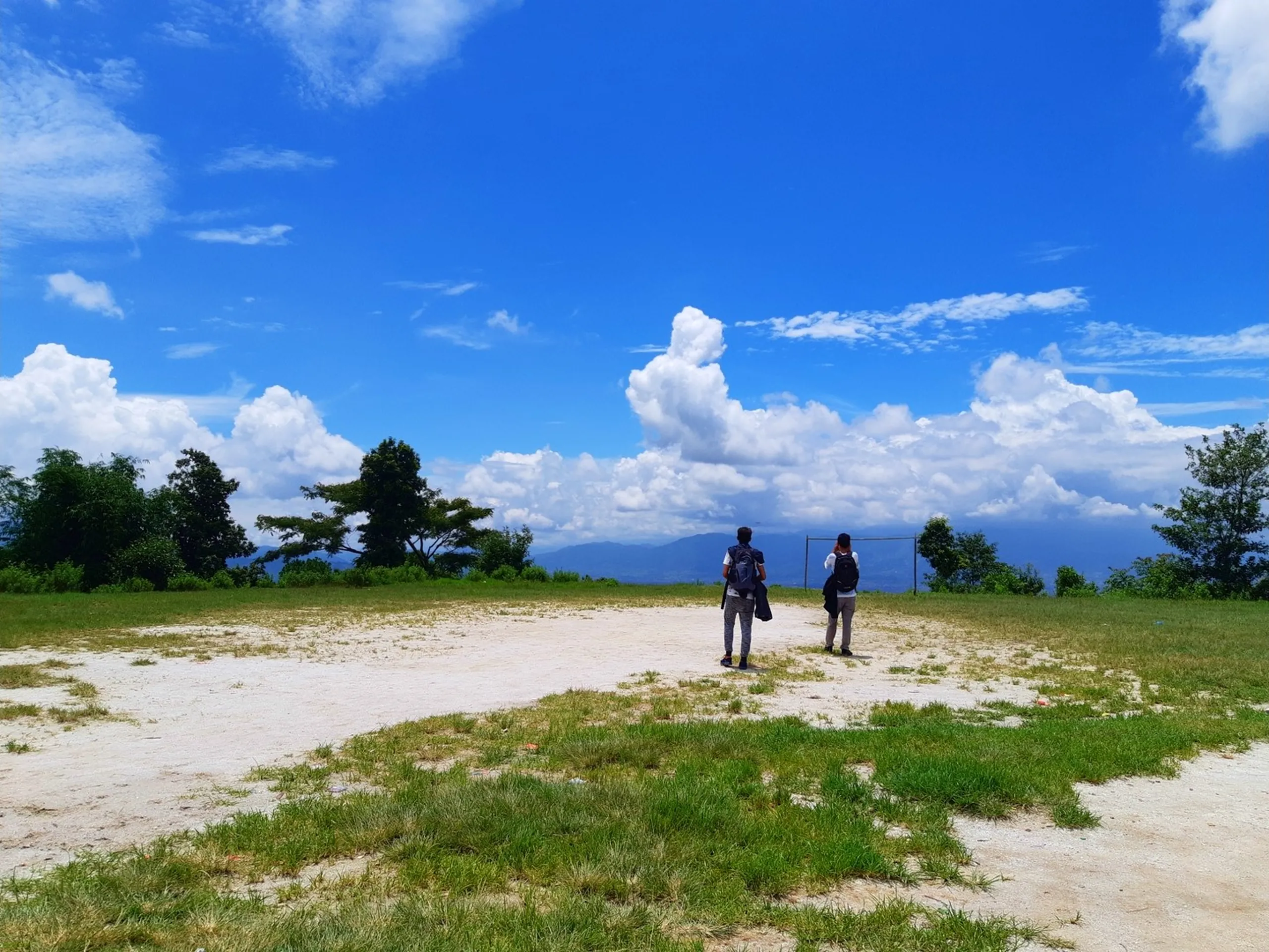 Two male hikers with backpacks standing on a wide open sandy hilltop clearing, facing away toward a sweeping panoramic view of distant blue mountain ranges and dramatic white cumulus clouds under vivid blue sky - Shivapuri Nagarjun National Park viewpoint Nepal hiking