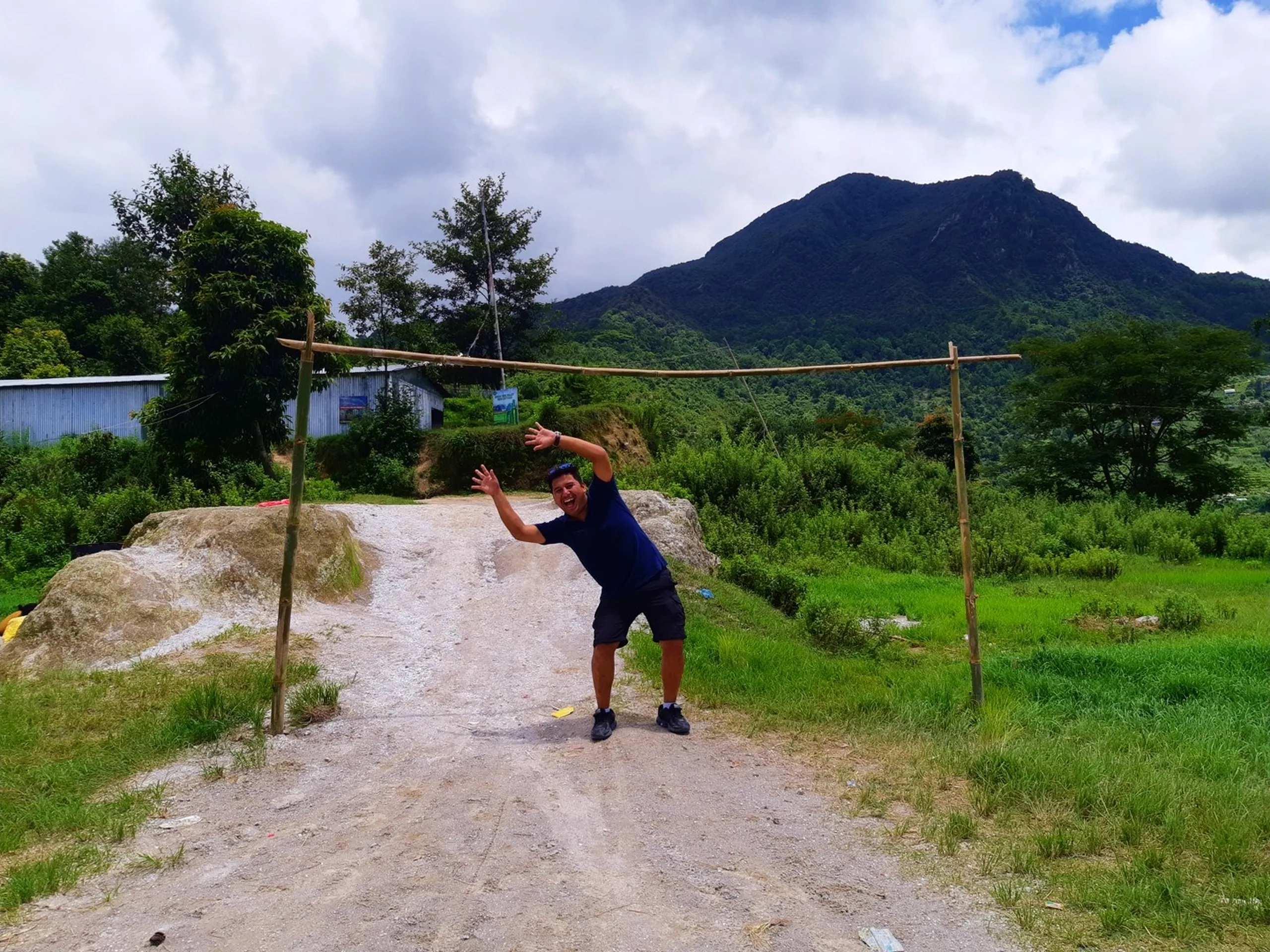 Energetic male hiker in blue t-shirt playfully posing with arms raised under a rustic bamboo gate structure on a gravel trail, dark forested mountain peak looming dramatically in the background - Nepal village trail adventure trekking day hike fun moment