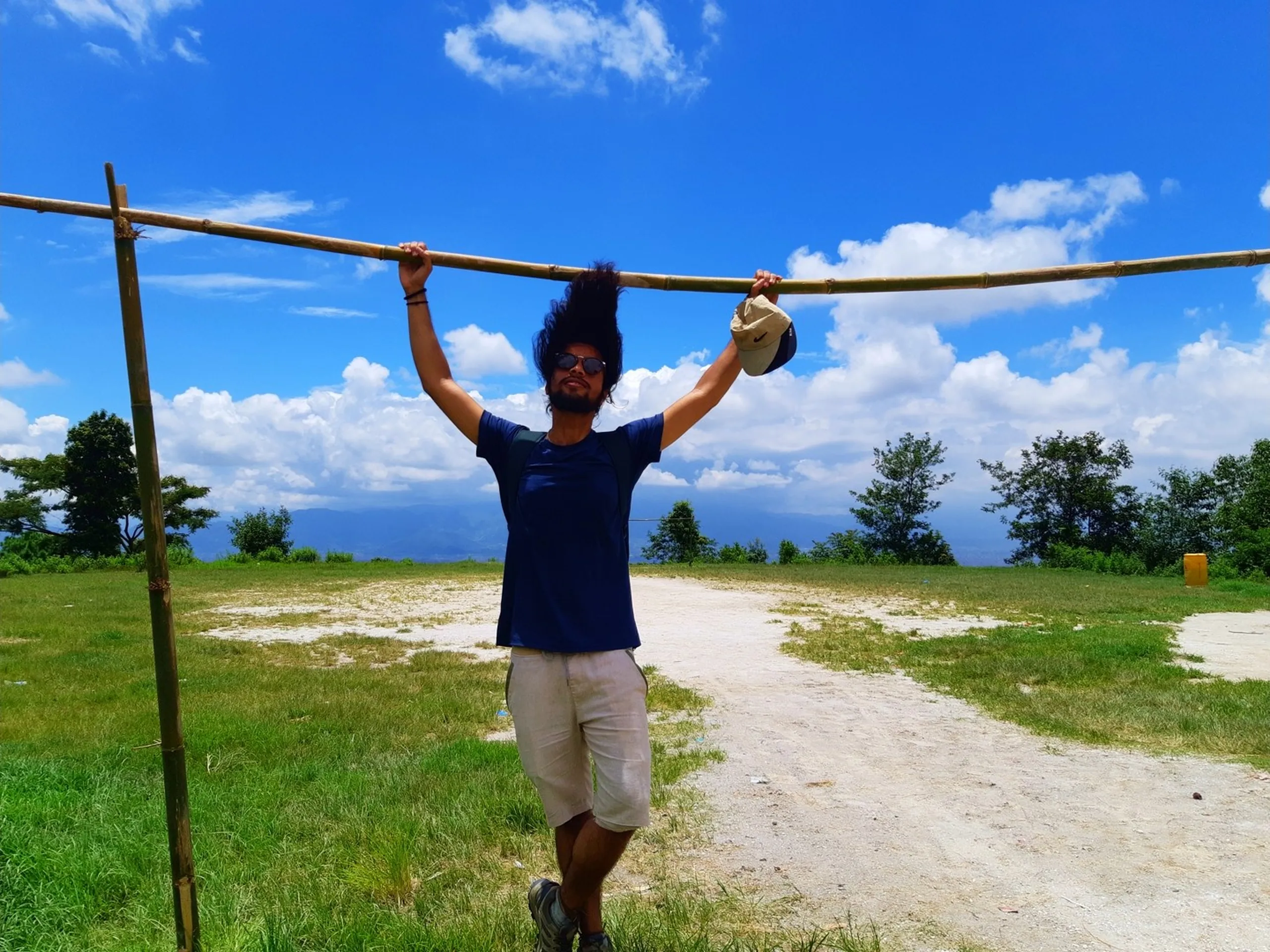 Young adventurous male traveler with wild curly hair hanging from a bamboo crossbar pole with arms stretched wide, sunglasses and cap in hand, bright blue sky with white clouds behind him, open grassy hilltop - Nepal hilltop trekking fun adventure travel Kathmandu